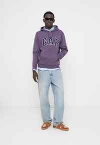 Sweat à capuche violet avec le logo "GAP", chemise à col bleu clair en dessous, jean bleu clair, et sandales marron, porté par une personne avec des lunettes de soleil.