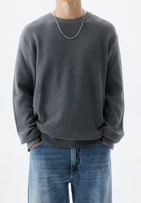 Grauer Waffelstrickpullover mit langen Ärmeln, lockerer Passform und umschlagenen Säumen, kombiniert mit verwaschenen blauen Jeans. Akzentuierter silberner Kettenanhänger.