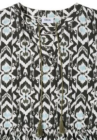 Blouse à motif géométrique noir et blanc avec des accents bleu clair, un col en V et des liens tressés à pompons suspendus au col.