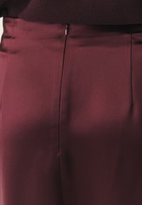 Burgundské kalhoty s hladkou, lesklou strukturou; mají středový záhyb a skrytý zip v pase pro elegantní vzhled.