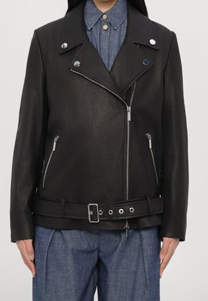 Personne portant une veste de motard en cuir noire avec des fermetures éclair argentées et des boutons sur une chemise bleue à col et un pantalon bleu.