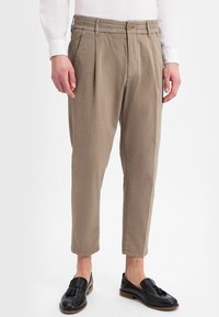 Man in beige, cropped, geplooide broek, een witte overhemd met lange mouwen en zwarte leren loafers met franje, staande tegen een witte achtergrond.