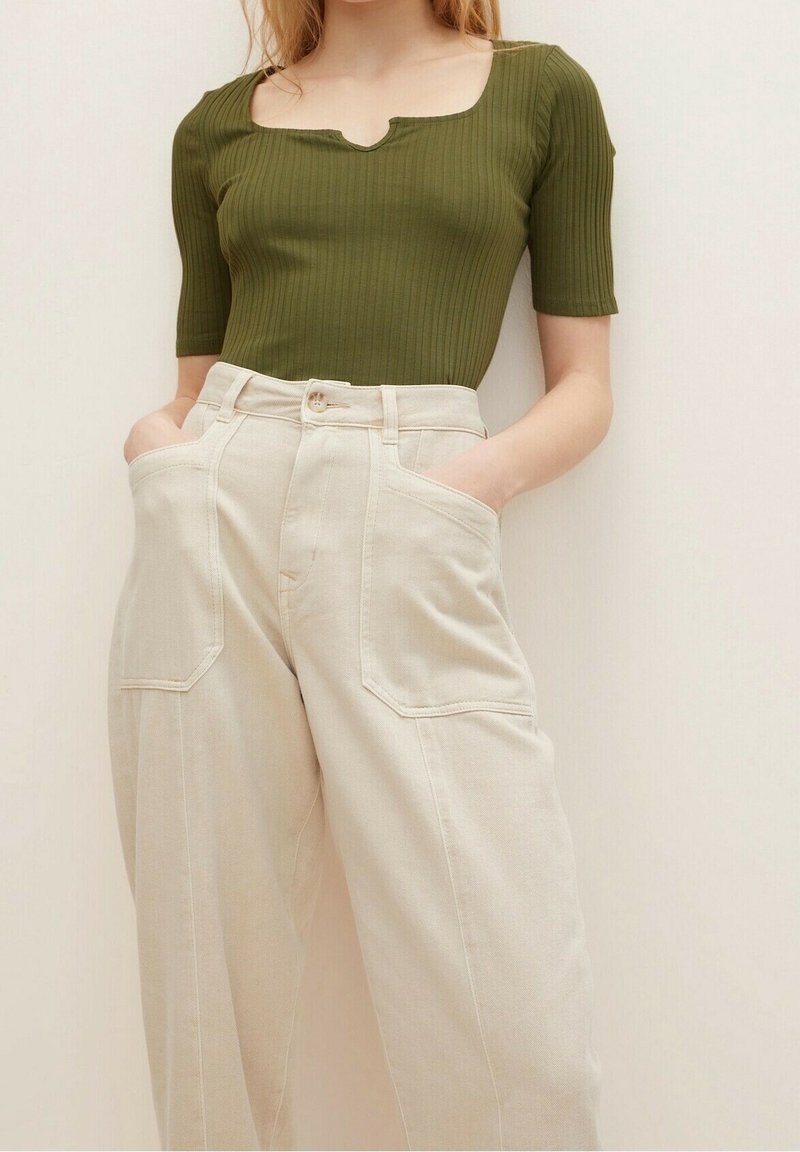Femme portant un haut côtelé à manches courtes couleur olive, rentré dans un pantalon crème taille haute, à jambes larges, avec de grandes poches frontales.