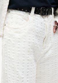 Pantaloni in denim bianco con texture, caratterizzati da un motivo a zigzag e dettagli sfilacciati; completati da una cintura nera con borchie a forma di stella.