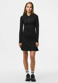 Zwarte poloshirt met lange mouwen en knopen, gecombineerd met een zwarte flared rok. Het model draagt zwarte sneakers en gestreepte zwart-witte sokken.