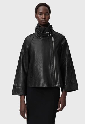 Vrouw met een loszittend zwart leren jasje met brede mouwen, een hoge kraag met gesp en een asymmetrische zilveren rits, gecombineerd met een zwarte rok.