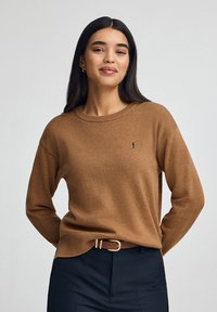 Brauner Pullover aus weichem Material, mit Rundhalsausschnitt, langen Ärmeln und einem dezenten Logo auf der linken Brust, kombiniert mit dunklen Hosen.