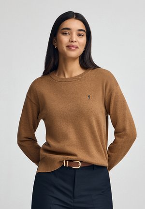 Pull marron en tissu doux, avec un col rond, des manches longues et un logo discret sur la poitrine gauche, porté avec un pantalon foncé.