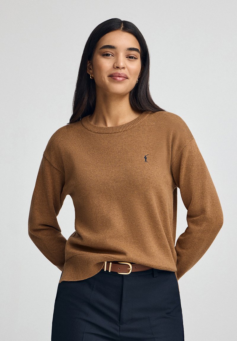 Brauner Pullover aus weichem Material, mit Rundhalsausschnitt, langen Ärmeln und einem dezenten Logo auf der linken Brust, kombiniert mit dunklen Hosen.