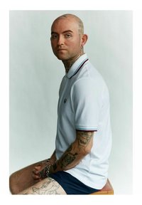 Polo bianca con colletto a strisce rosse e blu sottili, figura seduta con tatuaggi visibili, indossando pantaloni corti blu e accessori argento.