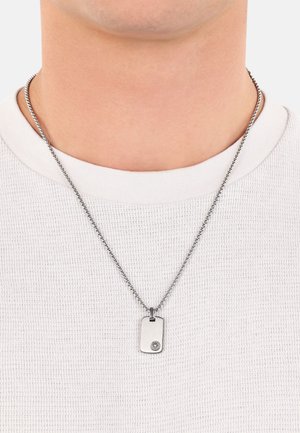 Silberfarbene Hundemarke als Anhänger an einer Kette, mit glatter Oberfläche und einem runden Emblem auf einer Seite. Wird über einem weißen Hemd getragen.