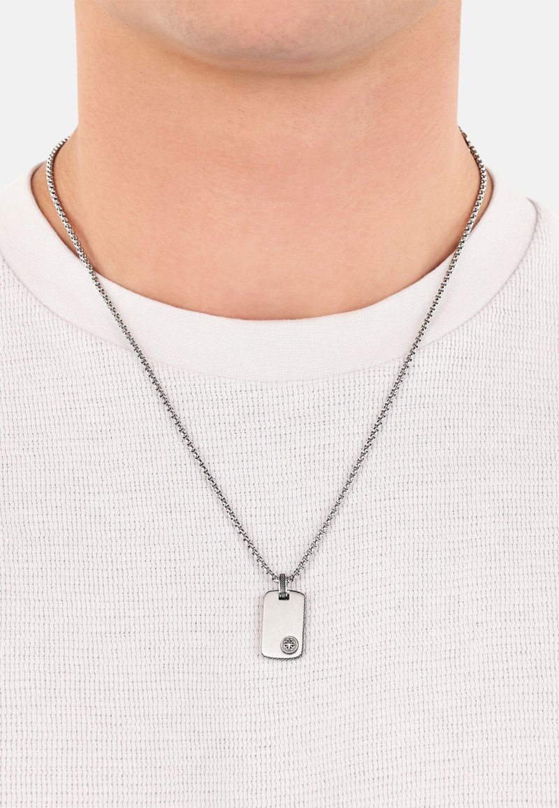 Collana con pendente a forma di dog tag in tonalità argento, su una catena, caratterizzata da una finitura liscia e un emblema circolare su un lato. Indossata sopra una maglietta bianca.