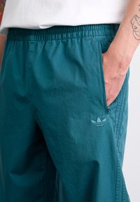 Pantalons de sport teal en tissu léger, avec une taille élastique, des poches latérales et un logo brodé sur la hanche.