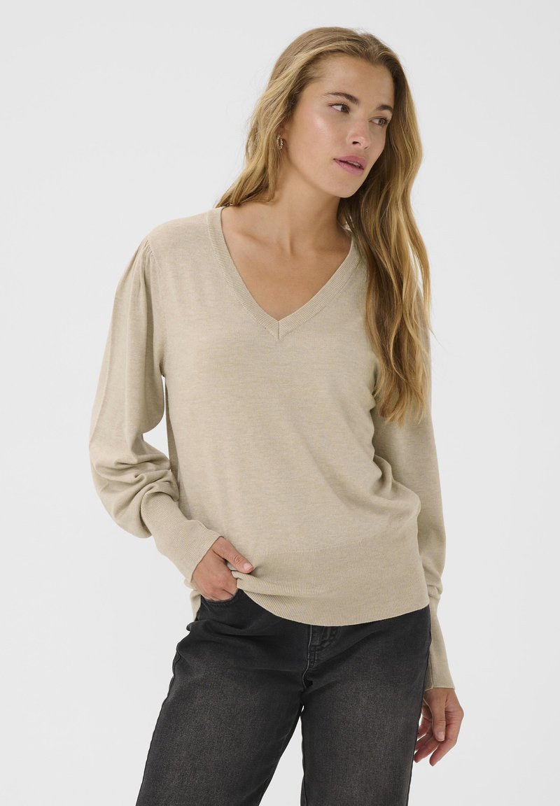 Maglione beige a V con maniche lunghe e leggermente a sbuffo. Tessuto a maglia morbida e polsini a coste, abbinato a jeans grigio scuro.