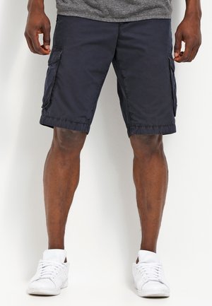 Mann trägt dunkelblaue Cargo-Shorts und weiße Sneaker, steht vor einem schlichten hellen Hintergrund.