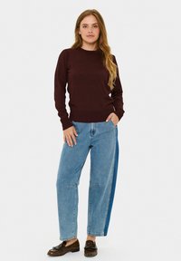 Burgundska pletena džemper s okruglim izrezom, uparen sa svijetloplavim širokim trapericama s bočnim džepovima i kontrastnim denim panelima. Smeđe mokasine.