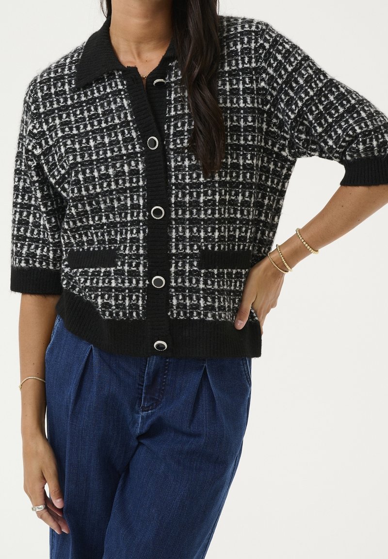 Femme portant un cardigan boutonné à carreaux noir et blanc avec bordure noire et un pantalon taille haute bleu foncé, bijoux au poignet et aux doigts.