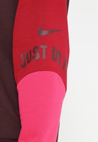 Sudadera de Nike con una mezcla de telas rojas, rosas y marrones. Incluye el texto "JUST DO IT" en gris oscuro en la sección roja.