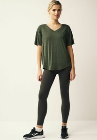 Olíva zöld, rövid ujjú, V nyakú felső, texturált felülettel, sötét leggings-szel párosítva. Fekete sportcipők fehér kiegészítőkkel.