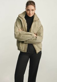 Khaki kurtka na zamek z wysokim kołnierzem, w oversize'owym kroju, z prążkowanymi mankietami; wyróżnia się teksturowanym materiałem i złotymi akcentami.