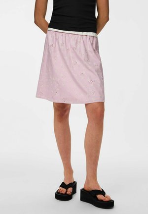 Žena v ružovej sukni po kolená s kvetinovými výrezmi, čiernom tielku bez rukávov a čiernych platformových sandáloch, stojaca s rukami za chrbtom.