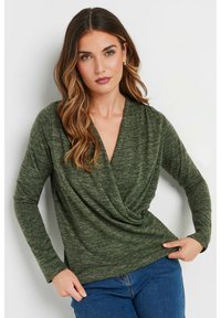 Långärmad, mörkgrön topp med crossover-design, tillverkad av ett mjukt, texturerat material. Kombinerad med blå denimjeans.