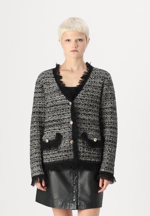 Cardigan de tweed en blanco y negro con tejido texturizado, bordes deshilachados, dos bolsillos frontales y botones dorados. Llevado sobre una blusa y falda negras.