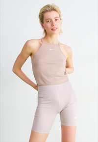 Top sportiv bej deschis și pantaloni scurți asortați, ambele din material elastic, având un mic logo alb Nike pe față.