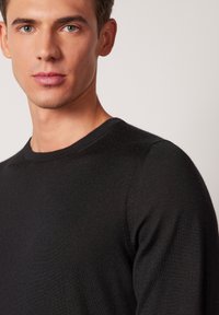 Svart stickad tröja med rund halsringning, mjukt material och figurnära passform. Fokuserar på överkroppen med naturligt ljus.