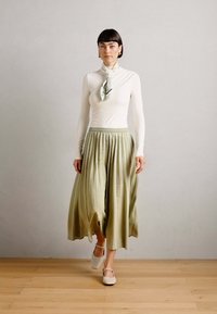 Lang, plissert oliven grønn skjørt parret med en tettsittende hvit topp. Modellen har på seg et silkeslips og kremfargede klipsteinsko. Minimalistisk design.