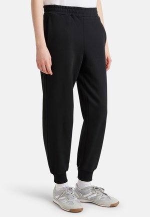 Zwarte sweatpants met een elastische tailleband, taps toelopende pijpen en ribbels aan de manchetten. Glad textuur en zijzakken. Draagt met grijze sneakers.