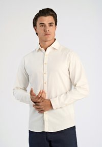 Camicia button-up a maniche lunghe crema chiaro in morbido tessuto, con colletto classico e bottoni marroni, abbinata a pantaloni scuri.