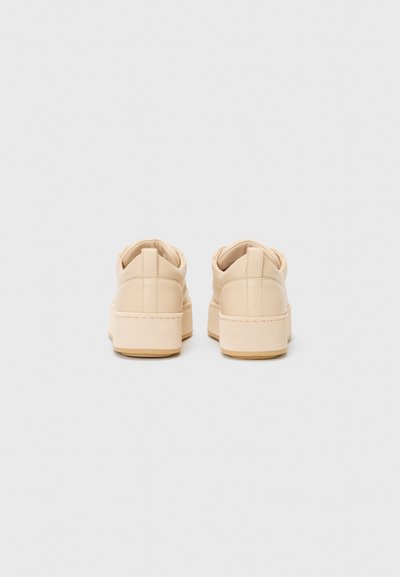 Baskets beige avec une texture lisse semblable à du cuir, à bout rond et une semelle épaisse et texturée. Design minimal, vue arrière sans lacets.