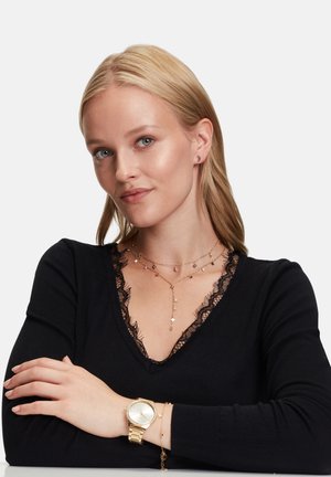 Goldene Uhr mit metallischem Armband, geschichtete Halsketten mit Stern- und Tropfenanhängern, schwarzes V-Ausschnitt-Top mit Spitzendetails.