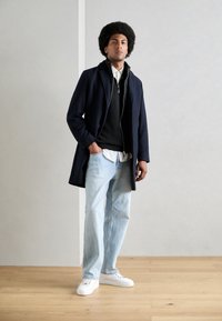 Abrigo navy sobre un suéter negro, camisa de cuello blanco, jeans de color azul claro y zapatillas blancas. Corte recto con una textura suave.