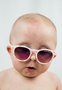 Pink and white patterned sunglasses with dark lenses worn by a baby. Plastic material, round shape, and visible brand on the frame.