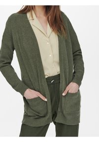 Olivově zelený cardigan s volným střihem, otevřeným předním dílem, žebrovanými rukávy a dvěma předními kapsami, vrstvený přes béžovou blůzu. Měkká textura.