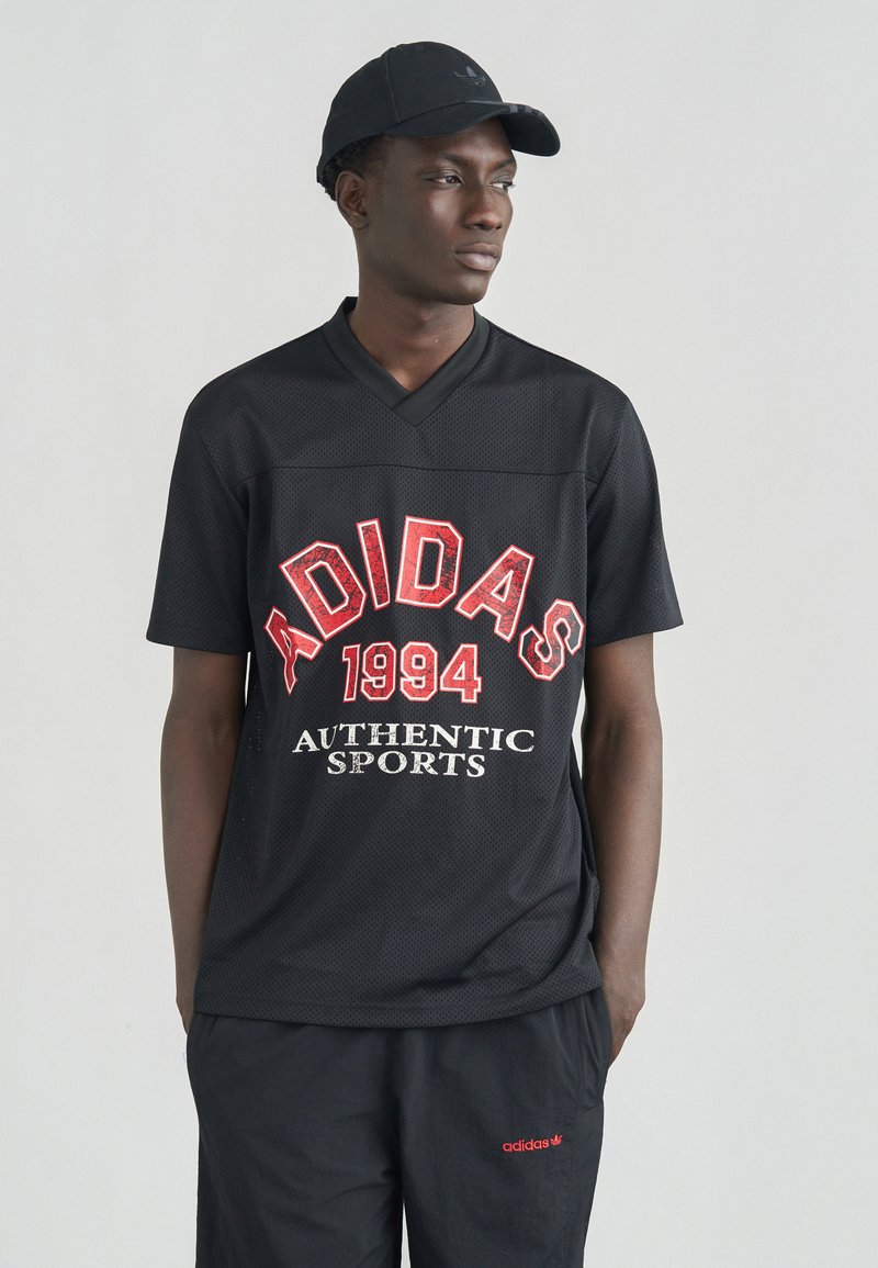 Νεαρός άνδρας με μαύρο καπέλο Adidas, μαύρο διαπνέον T-shirt με το κείμενο "Adidas 1994 Authentic Sports" και μαύρο παντελόνι Adidas με κόκκινο λογότυπο.