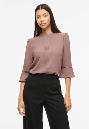 Femme aux longs cheveux foncés porte un chemisier mauve avec des plis et une bordure en dentelle aux manches cloche, associé à un pantalon noir taille haute, debout sur un fond neutre.