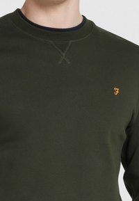 Olivově zelená mikina s kulatým výstřihem, s malým vyšitým oranžovým logem na levé hrudi a texturovanými švy.