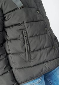 Siva puffer jakna s ušivenim dizajnom, teksturiranom tkaninom, jednim zatvorenim džepom na lijevoj strani i tamnim remenom na ramenu.