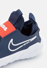 Baskets de sport bleu marine avec une sangle élastique rouge et un logo Nike blanc. Elles disposent d'un dessus en synthétique lisse et d'une semelle blanche rembourrée.
