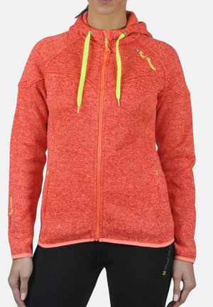 Femme portant une veste à capuche zippée orange vif avec des cordons jaunes et un pantalon noir, montrée de la nuque aux hanches.