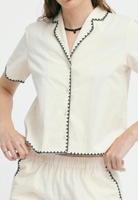 Top corto color beige chiaro con rifiniture a zigzag nere, chiusura frontale con bottoni e vestibilità comoda, abbinato a pantaloni corti con vita elastica dello stesso colore.