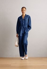 Combinaison en denim à manches longues, taille cintrée, deux poches poitrine et coupe décontractée. Portée avec des chaussures blanches et tenant un petit sac à main.