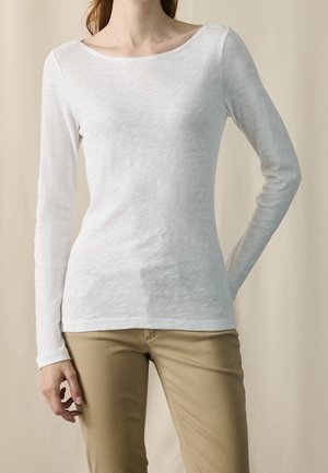 Frau trägt ein schlichtes weißes Langarmshirt und beige Hose, steht mit einer Hand auf der Hüfte und der anderen entspannt.