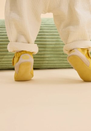 Geel baby schoenen met grijze accenten en een gestructureerde zool, gecombineerd met opgerolde crèmekleurige katoenen broeken, gelegen op een groene ribbeloppervlakte.