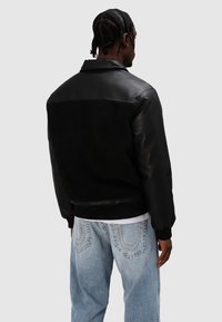 Bomberka z czarnej skóry i kożucha z prążkowanymi mankietami i ściągaczem, z gładkim przodem i fakturowanym tyłem. Noszona na jasnoniebieskich dżinsach.