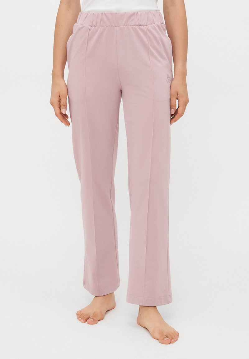 Femme portant des pantalons de lounge droits avec une taille élastique rose clair, se tenant pieds nus contre un fond blanc uni.