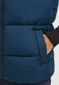 Jūrinis pūkinis liemuo su blizgiu nailono išoriniu paviršiumi, su užtrauktuku priekyje, šoniniais kišeniais ir įtemptais elastiniais rankogaliais papildomam komfortui.